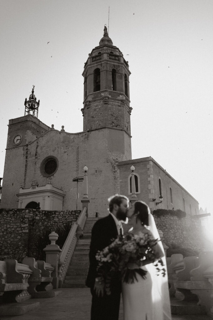 Bride and groom kissing in front of a historic stone church during Barcelona elopement. The scene is black and white with a romantic, timeless mood.