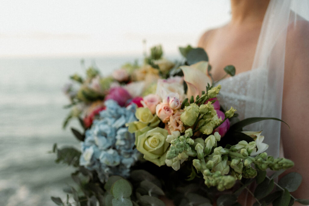 Bride holds colorful bouquet of roses and hydrangeas, with ocean in soft focus background. Mood is serene and romantic.