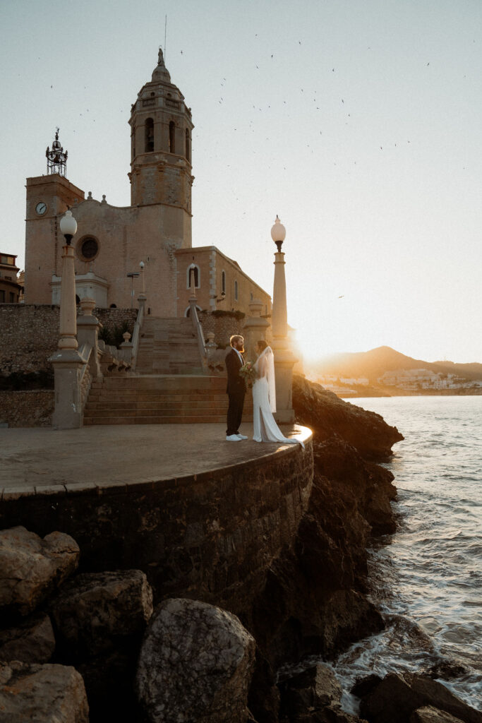 Bride and groom stand by a seaside church during Barcelona elopement, with the ocean and mountains in the background, creating a romantic scene.