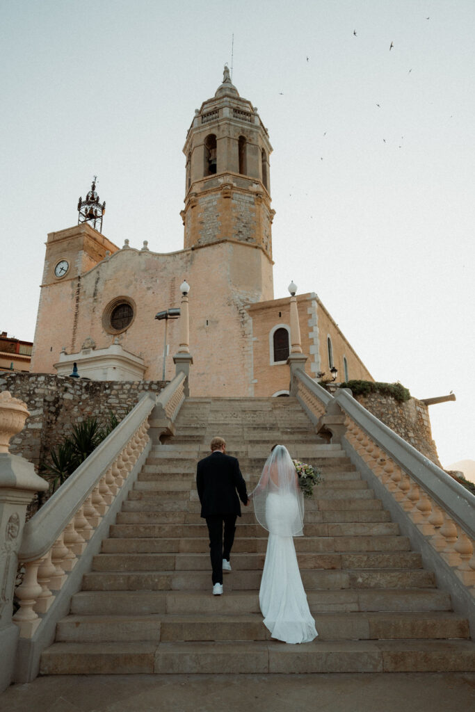 Bride and groom ascend stone steps towards an old Barcelona church during elopement. Bride holds bouquet. Evening sky with birds creates a serene, romantic mood.