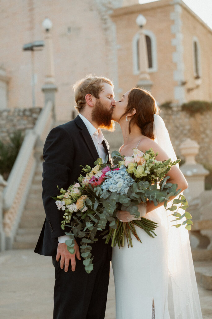 Bride and groom kissing, holding a colorful bouquet during their elopement. They're in front of a stone building in Barcelona, illuminated by warm, golden sunlight.