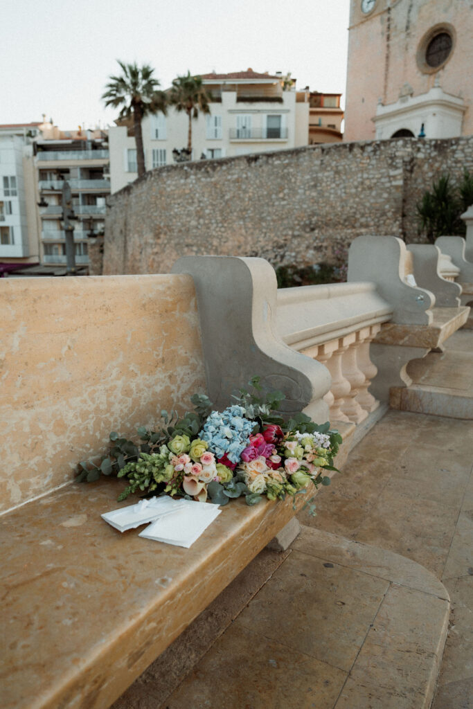 Elopement bouquet of colorful flowers on a stone bench with papers, set against a backdrop of an old wall and buildings of Barcelona. Peaceful and serene mood.