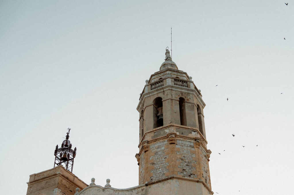 Stone tower with clock and bell, birds flying around under a clear, pale sky. Weathered Barcelona architecture evokes a serene, historic mood.