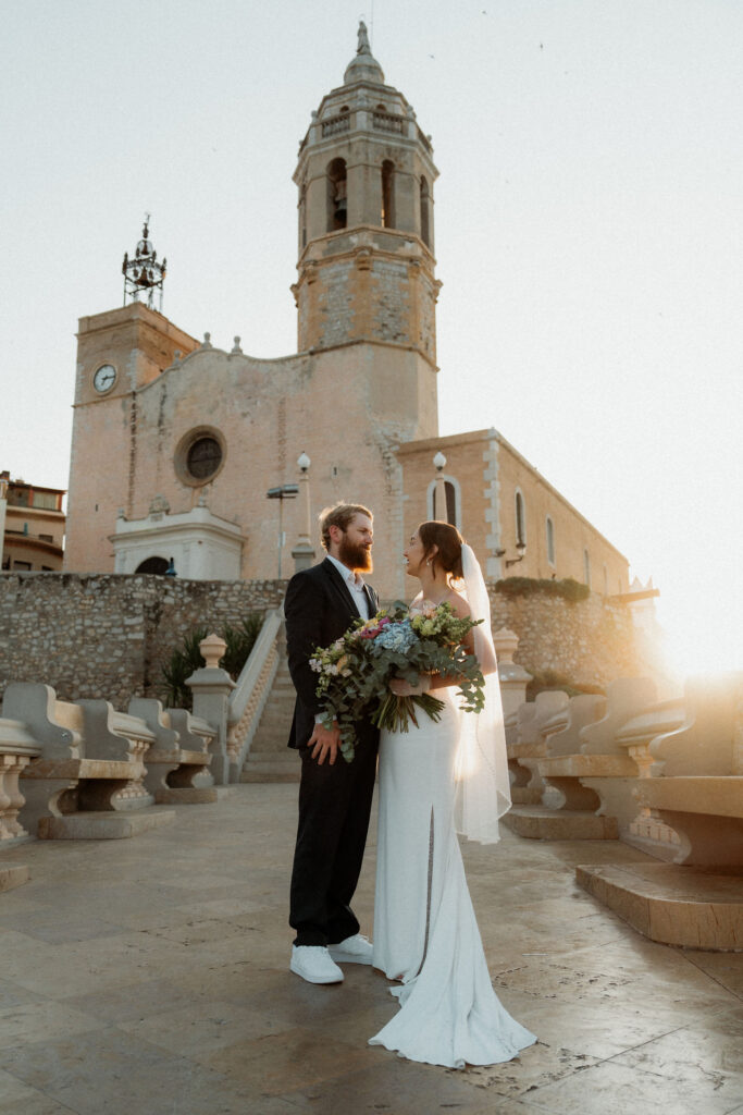 Bride and groom smiling under a clear sky, holding an elopement bouquet. Sunlight and an old brick church set the mood with warm tones in Barcelona.