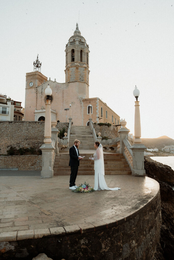 Bride and groom exchange vows on stone platform in Barcelona by historic church at sunset, with bouquet on ground. Calm, romantic setting.