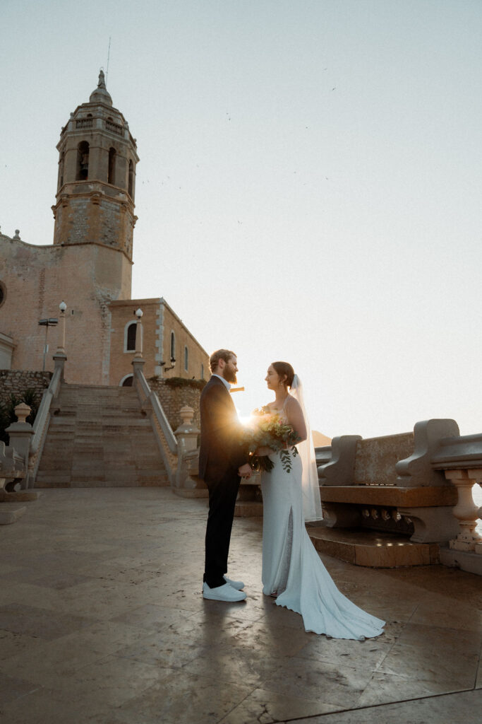 Bride and groom stand facing each other at sunset, holding flowers. Historic church tower in Barcelona. Romantic and serene mood.