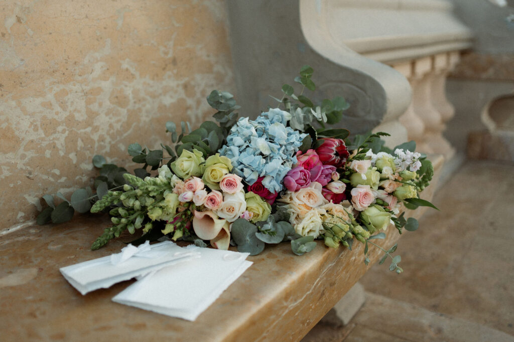 Colorful elopement bouquet with roses and hydrangeas on a stone bench. Beside it, white envelopes lie neatly stacked. Background shows ornate stonework.