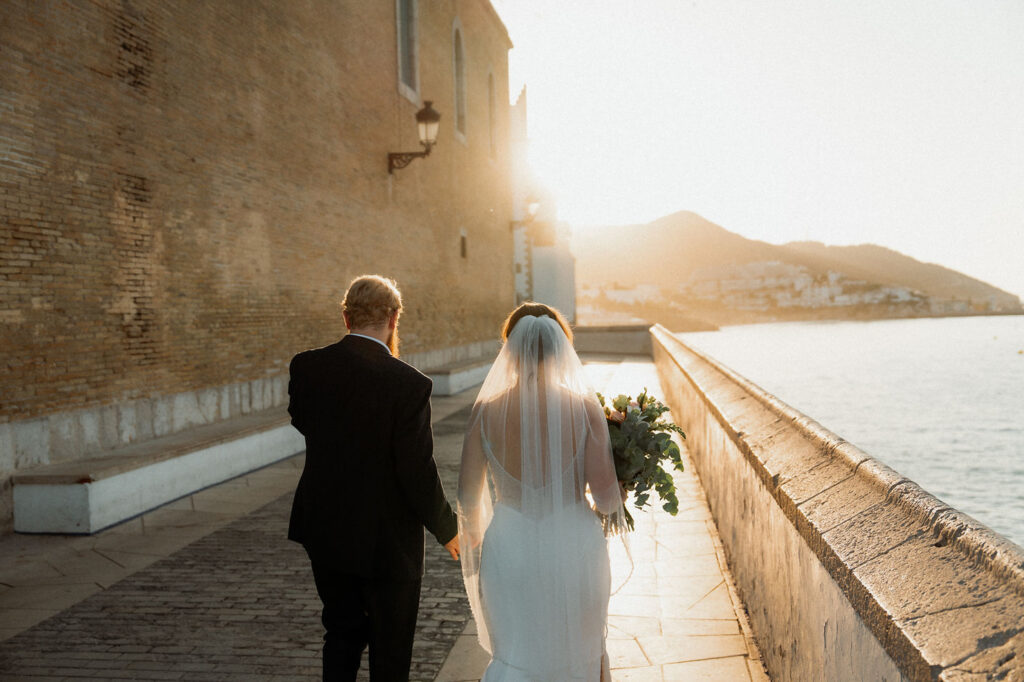 Bride and groom walk hand in hand by a sunlit seaside wall in Barcelona during wedding.