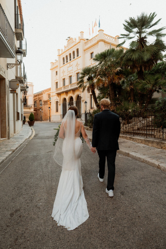 Bride and groom holding hands walk down a quaint street in Barcelona during elopement. She wears a white dress and veil; he's in a dark suit. Elegant building and palms ahead.