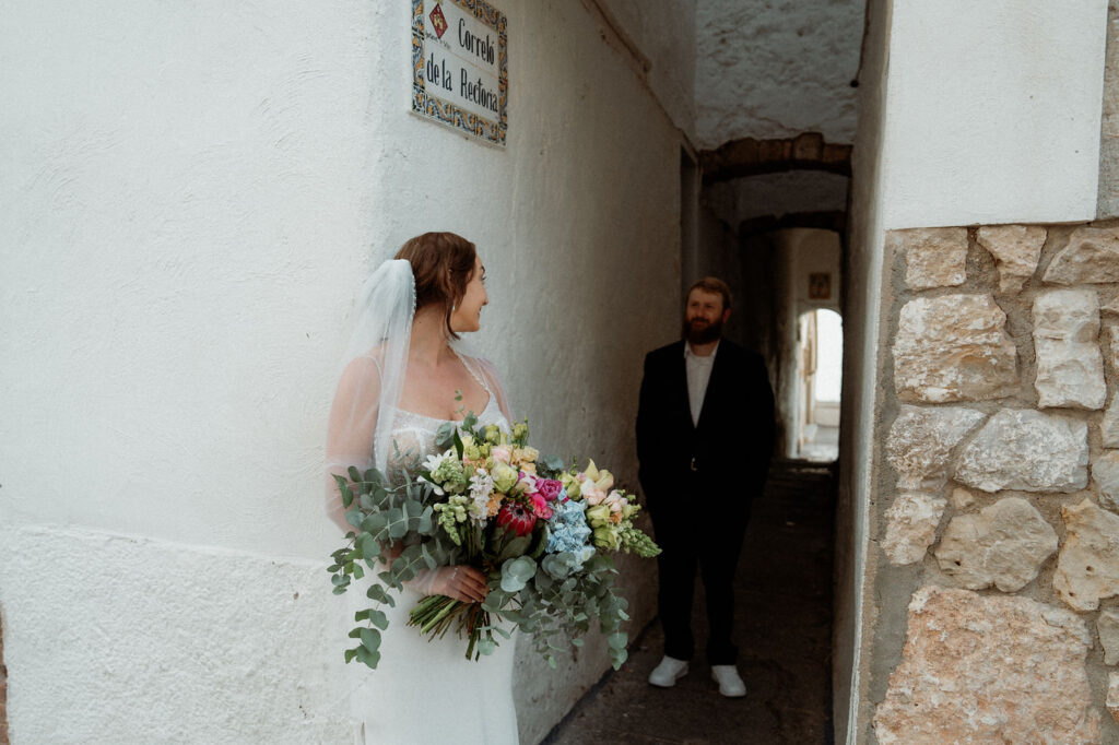 Bride with colorful bouquet smiles at a man in a narrow stone alley during Barcelona wedding.