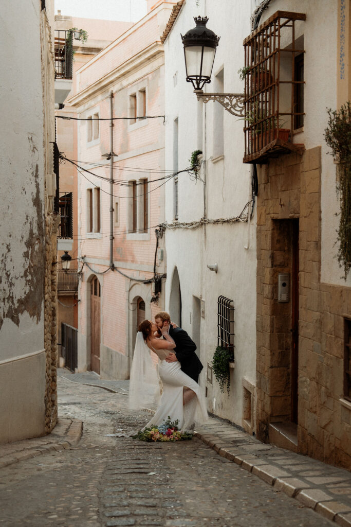 Bride and groom kissing on a narrow cobblestone street in spain. Bride in a white dress; buildings with pastel walls in the background; romantic mood.