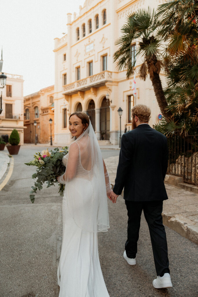 Bride in white dress and veil holding elopement bouquet, smiling back, walks hand in hand with groom in Barcelona
