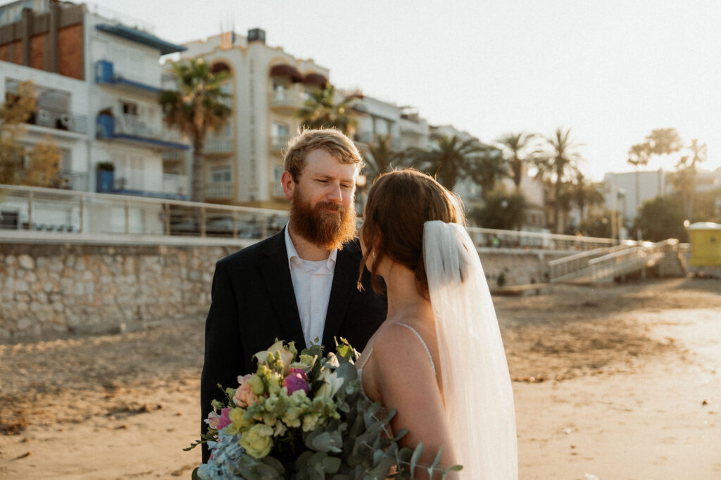 Bride and groom facing each other on a beach at sunset, bride holding a bouquet. Background of palm trees and buildings, creating a romantic scene.