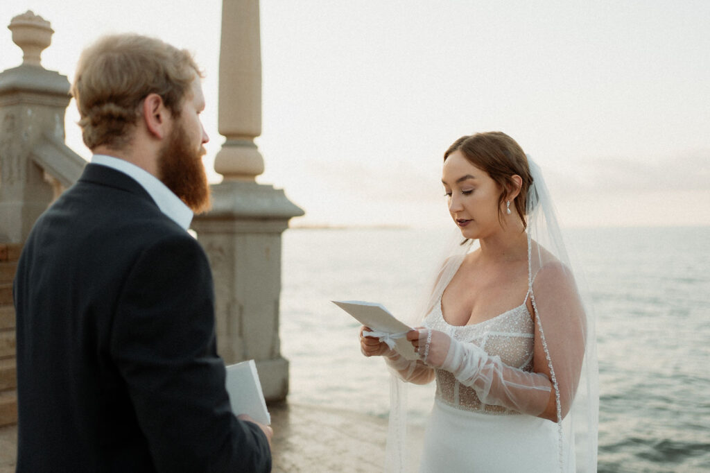 Bride and groom exchange vows by the Barcelona sea at sunset. The bride wears a white dress and veil, holding a card. Stone pillar and steps nearby.