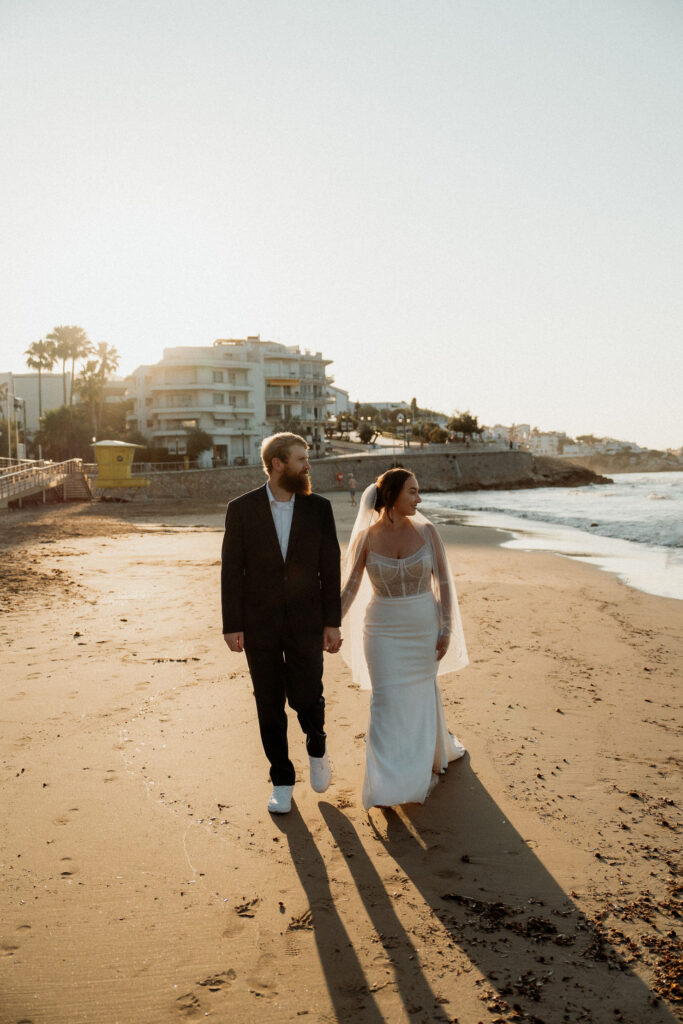 Bride and groom walking hand in hand on a sunny beach in Barcelona, with buildings and palm trees in the background. The mood is romantic and serene.