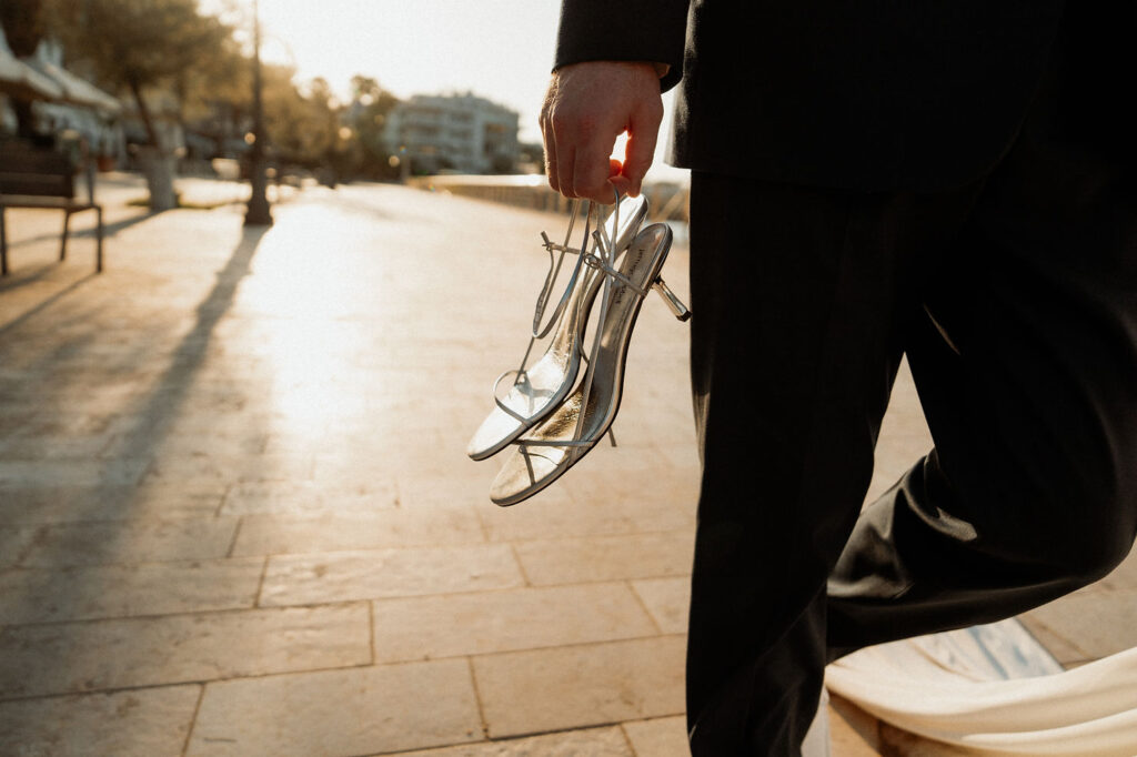 A person in a suit holds silver high heels while walking on a sunlit street in Barcelona after elopement.