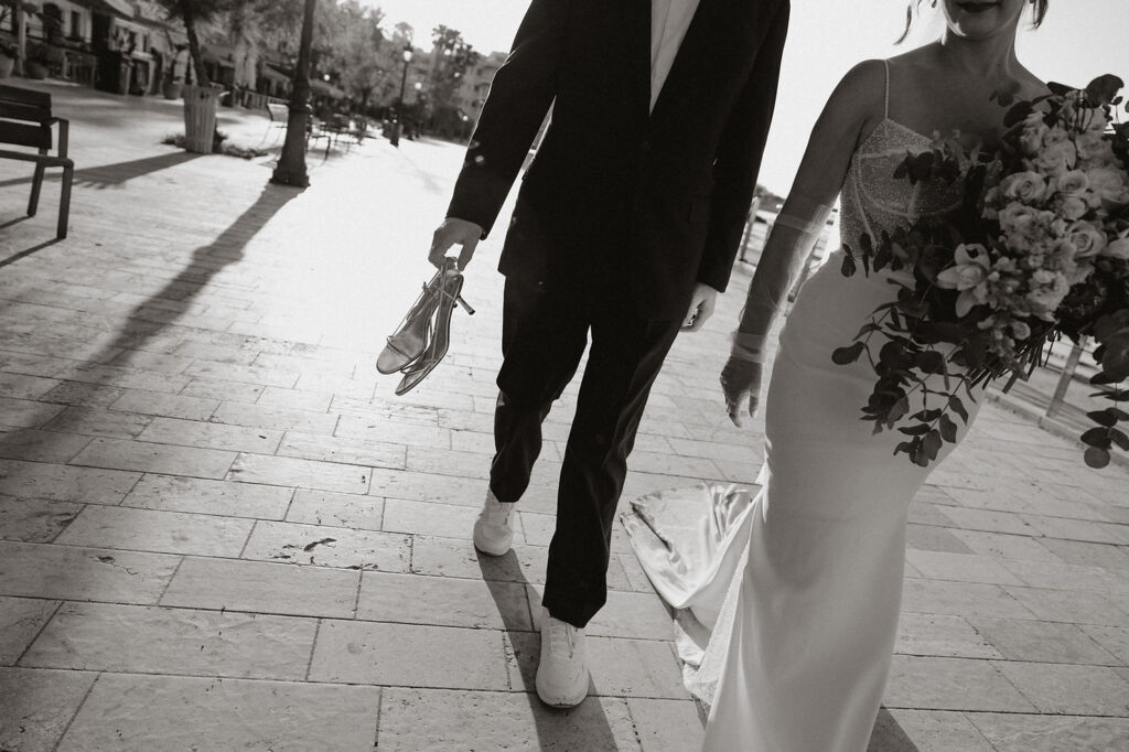 Bride and groom walking on a sunlit path; groom holds shoes, bride carries wedding bouquet.