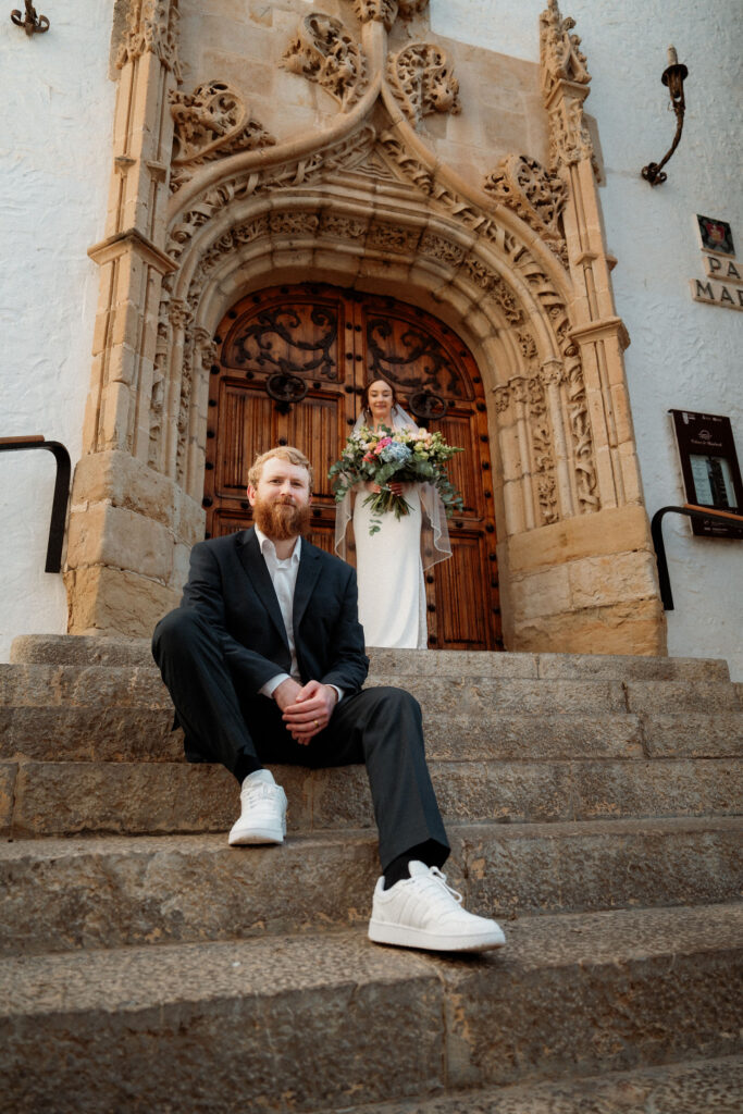 groom sits on stone steps, bride with bouquet stands behind under ornate arch during elopement photos. Historic building in Barcelona, elegant mood.