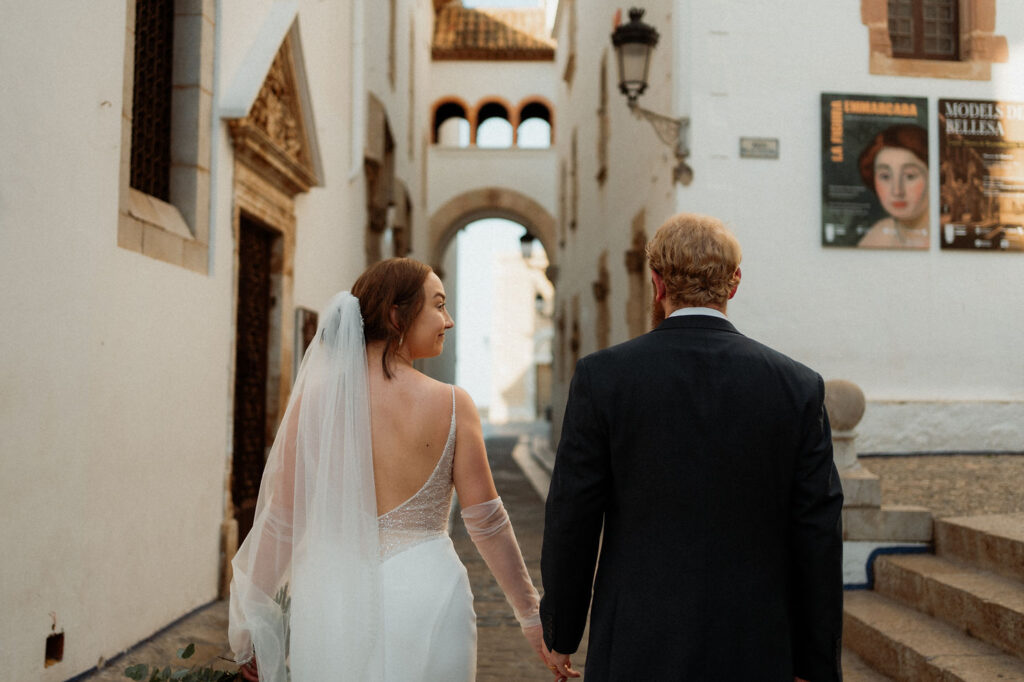 Bride walks hand-in-hand with groom in dark suit down a sunny, narrow street lined with buildings and posters in Barcelona