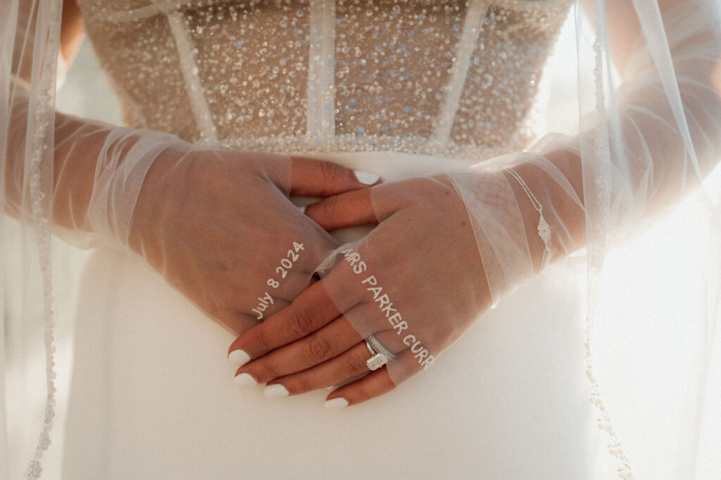 Bride's gloved hands clasped, wearing a ring. Veil and dress have sequins. Text on gloves reads: July 8, 2024, Mrs. Parker Curry.