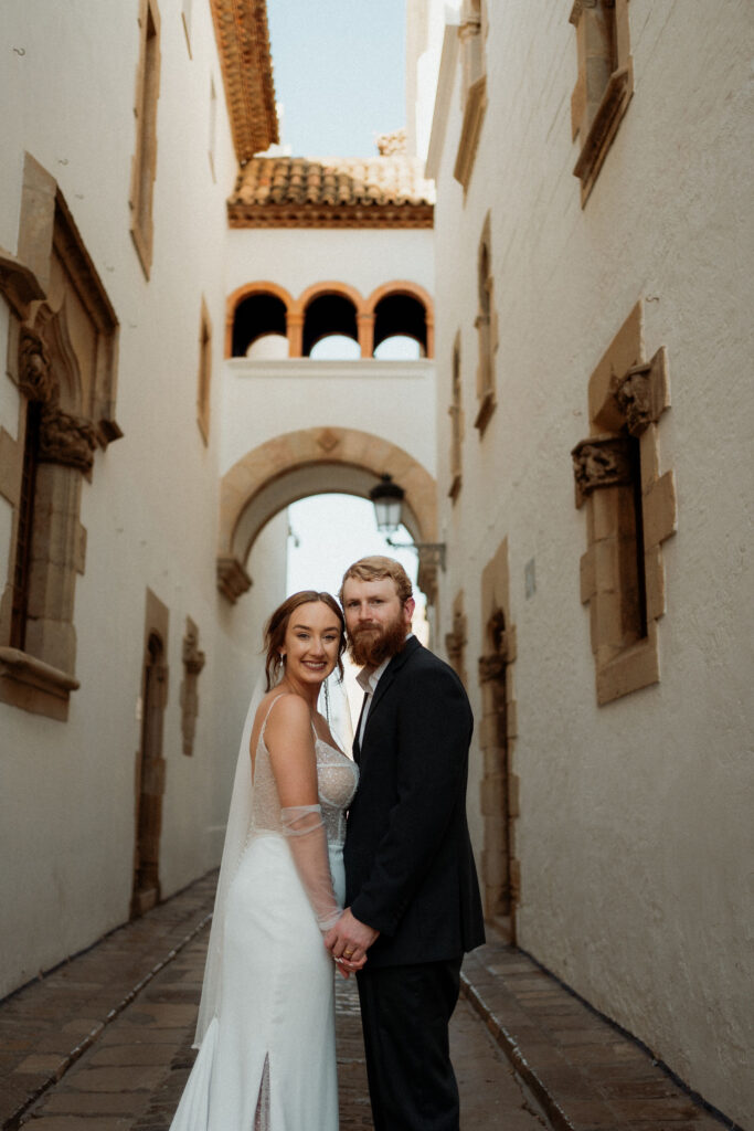 Bride and groom hold hands in a narrow alley with stone walls and arches during Barcelona wedding