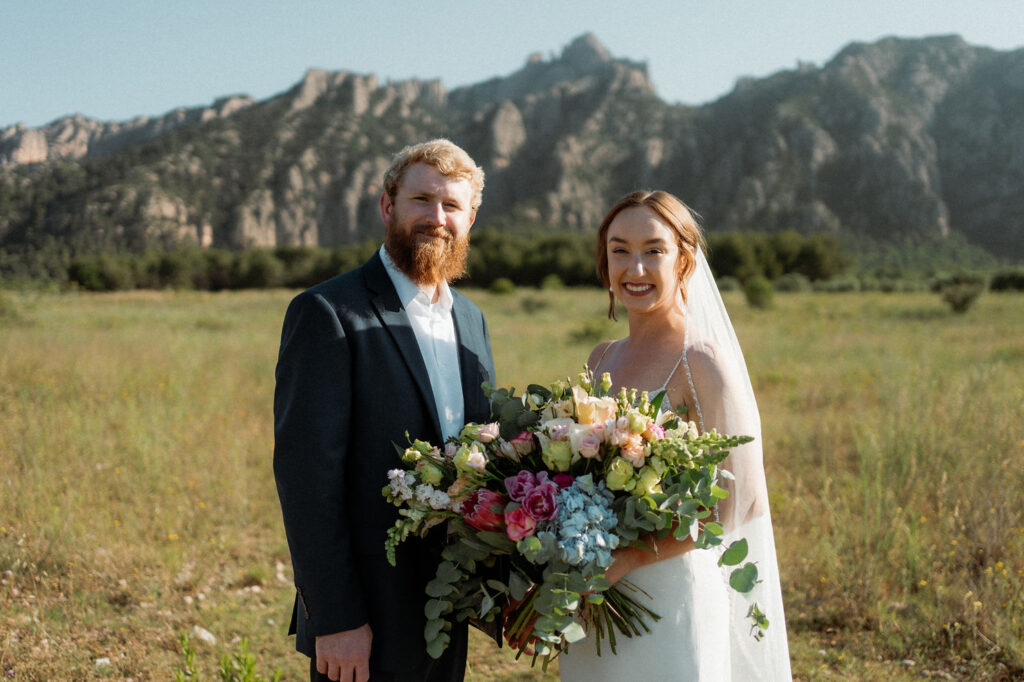 Bride and groom smiling in front of spanish mountains, she holds a colorful elopement bouquet. Sunny outdoor setting with greenery and clear sky.