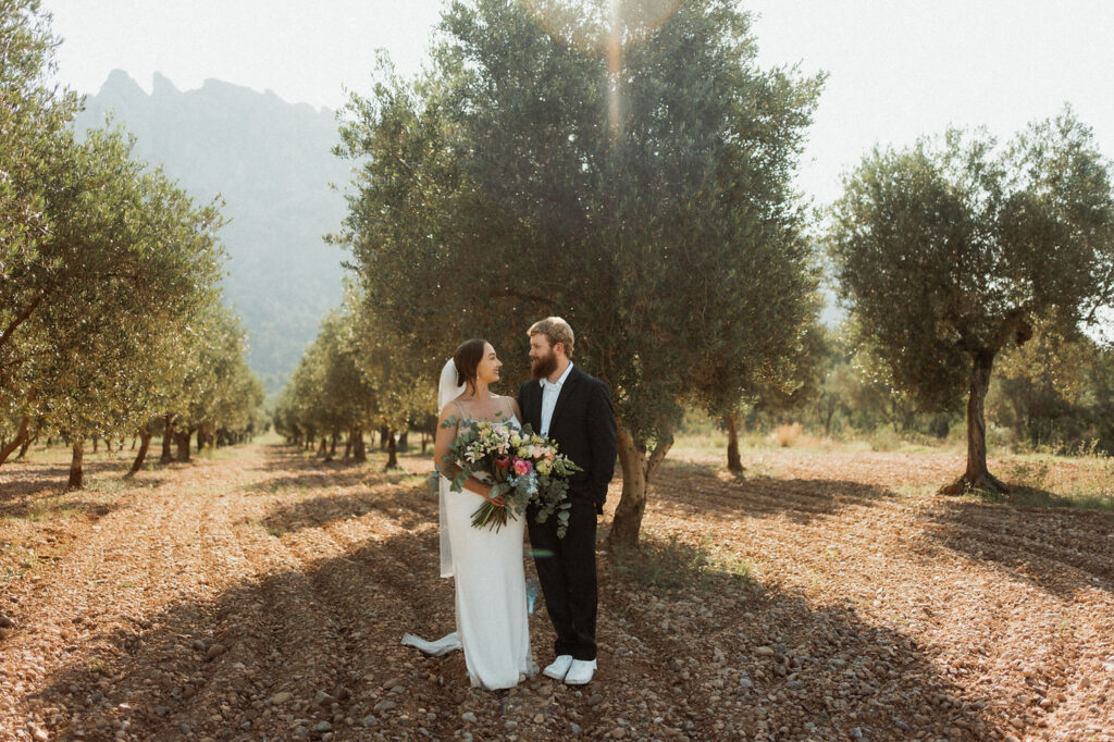 Bride and groom stand smiling in an olive grove, with mountains in the background. She holds a bouquet; sunlight filters through trees.