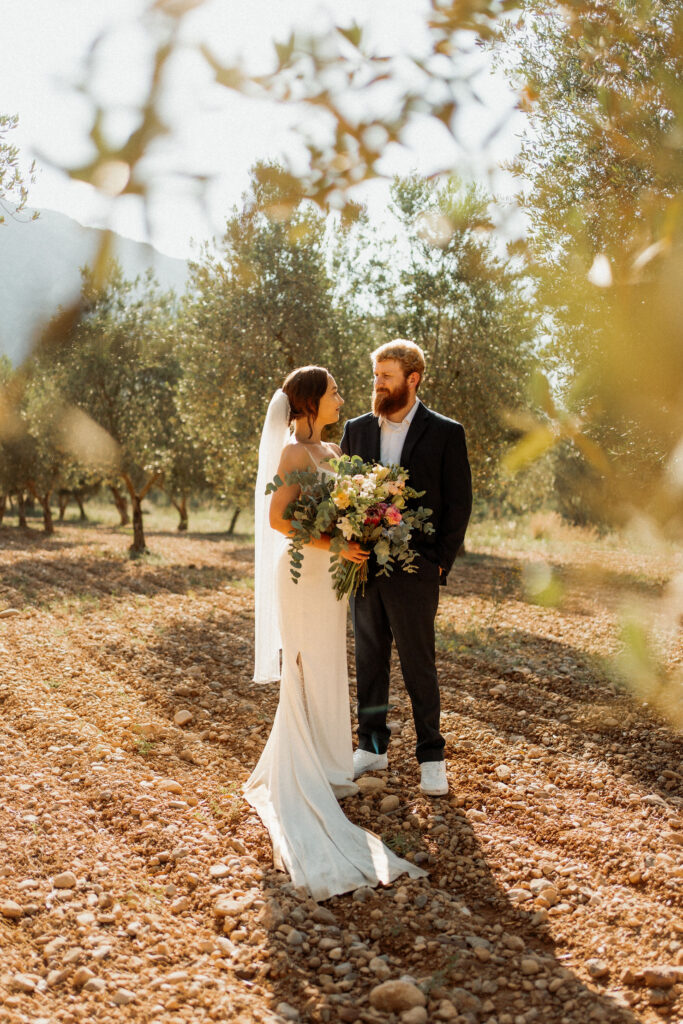 Bride and groom stand lovingly in a sunny olive grove. She holds a colorful elopement bouquet, wearing a white dress and veil. Romantic atmosphere.