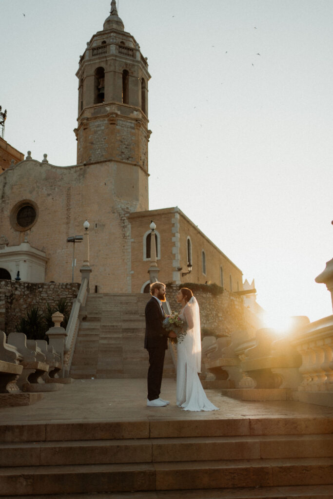 Bride and groom stand on stone steps, exchanging vows at sunset in Barcelona. Background features a historic tower, creating a romantic ambiance.
