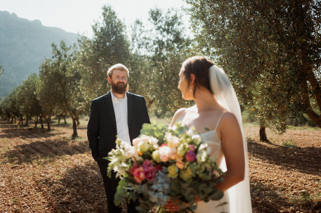 Bride with veil holds colorful bouquet, smiling at groom in suit amidst sunlit olive trees and mountains, conveying a joyful mood.