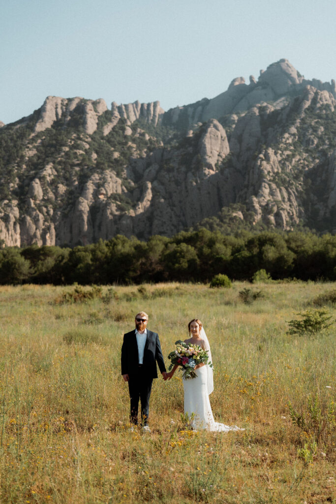 Bride and groom stand in a sunny meadow outside of Barcelona holding hands, with mountains in the background. She holds a colorful bouquet. Romantic mood.