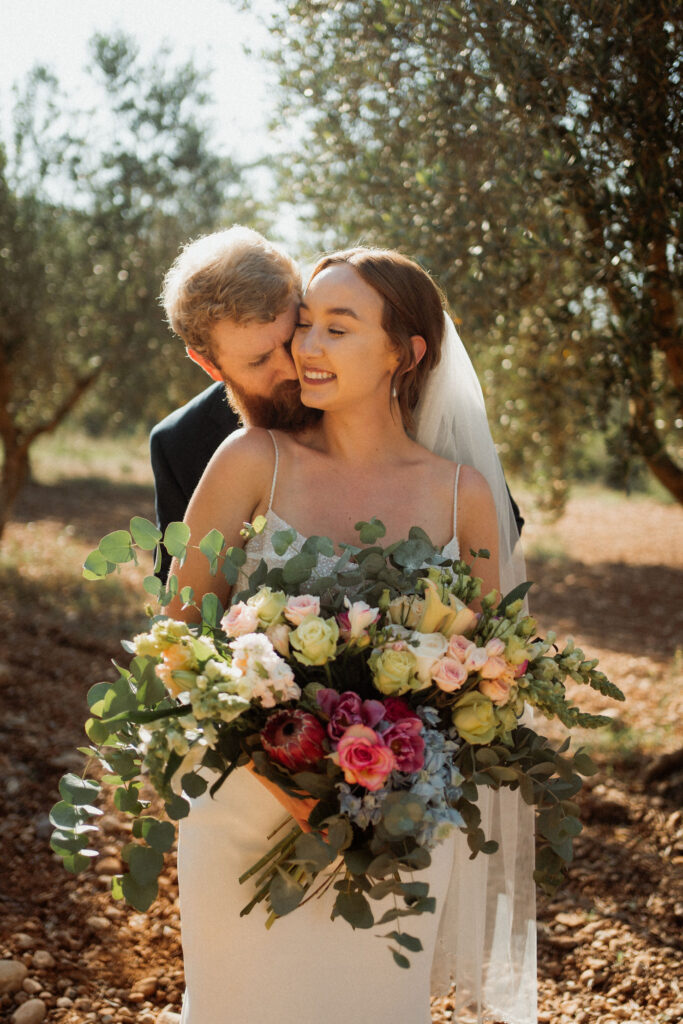 Bride holding vibrant bouquet, groom affectionately nuzzles her from behind. Sunlit olive grove creates a serene backdrop.