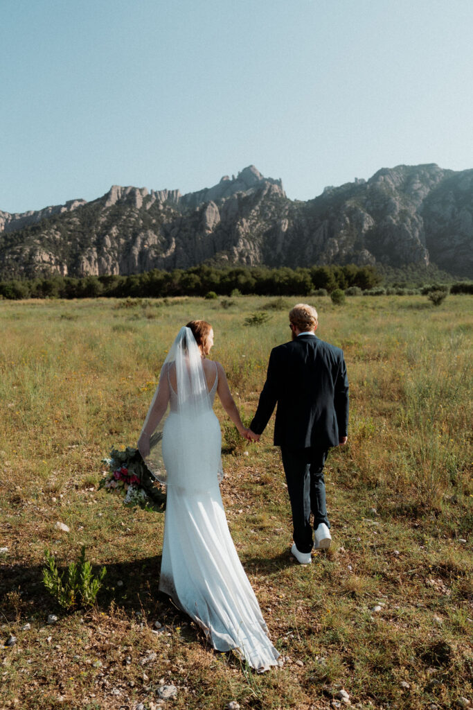 Bride and groom holding hands, walking on grassy field with mountain backdrop during Barcelona elopement