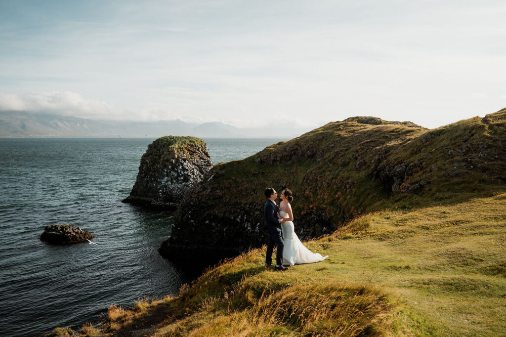 during iceland elopement at snæfellsnes peninsula bride and groom embrace on a grassy cliff by the ocean, with rocky formations and mountains in the distance under a clear sky.