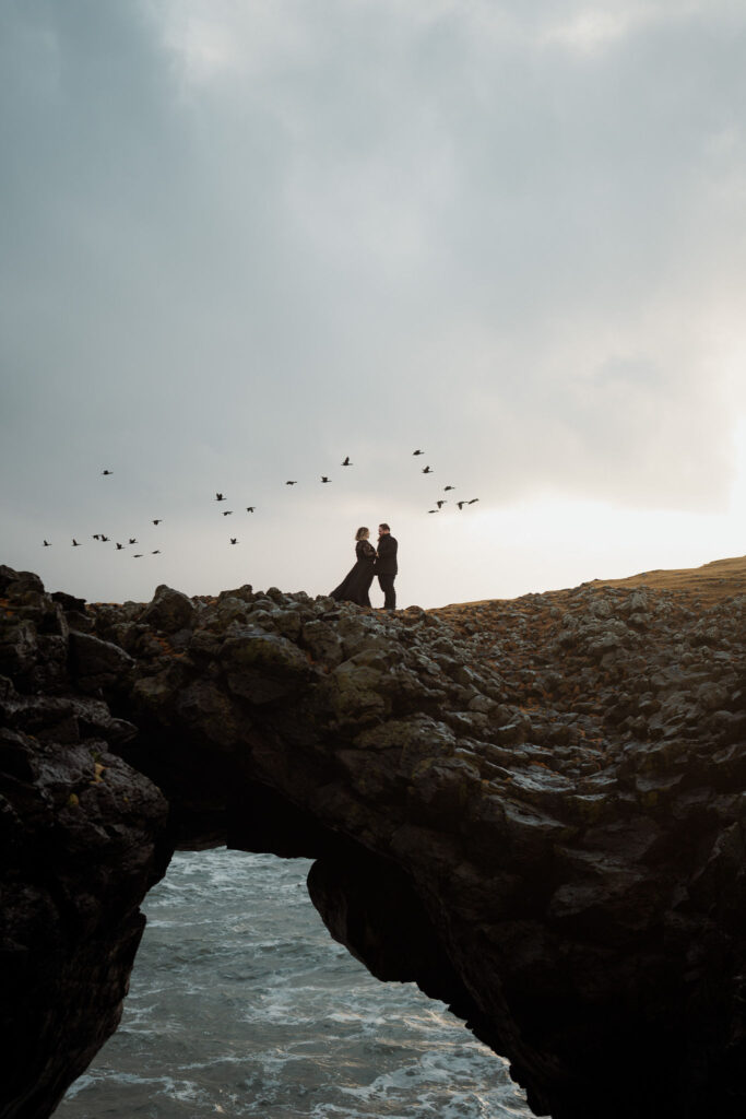 A couple stands on a rocky arch with birds flying above, set against a cloudy sky and ocean background, creating a serene and romantic mood for their iceland elopement at snæfellsnes peninsula