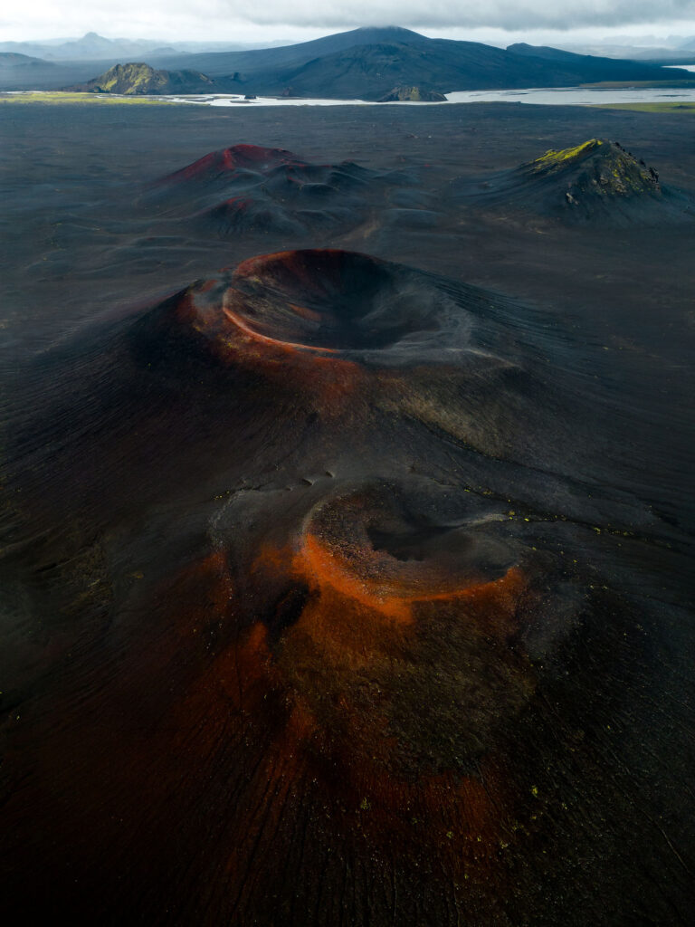 best place to elope in iceland: Fjallabak Nature Reserve. Aerial view of volcanic craters with red and orange hues, surrounded by dark terrain. Mountains and a lake in the background under cloudy skies.