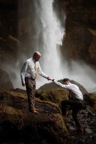 after deciding what it costs to elope in iceland, LGBTQ+ couple climbs on rock in front of waterfall