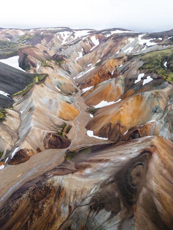 Aerial view of colorful, snow-speckled mountains and valleys with vibrant hues of orange, brown, and green, creating a dramatic landscape at Fjallabak Nature Reserve.
