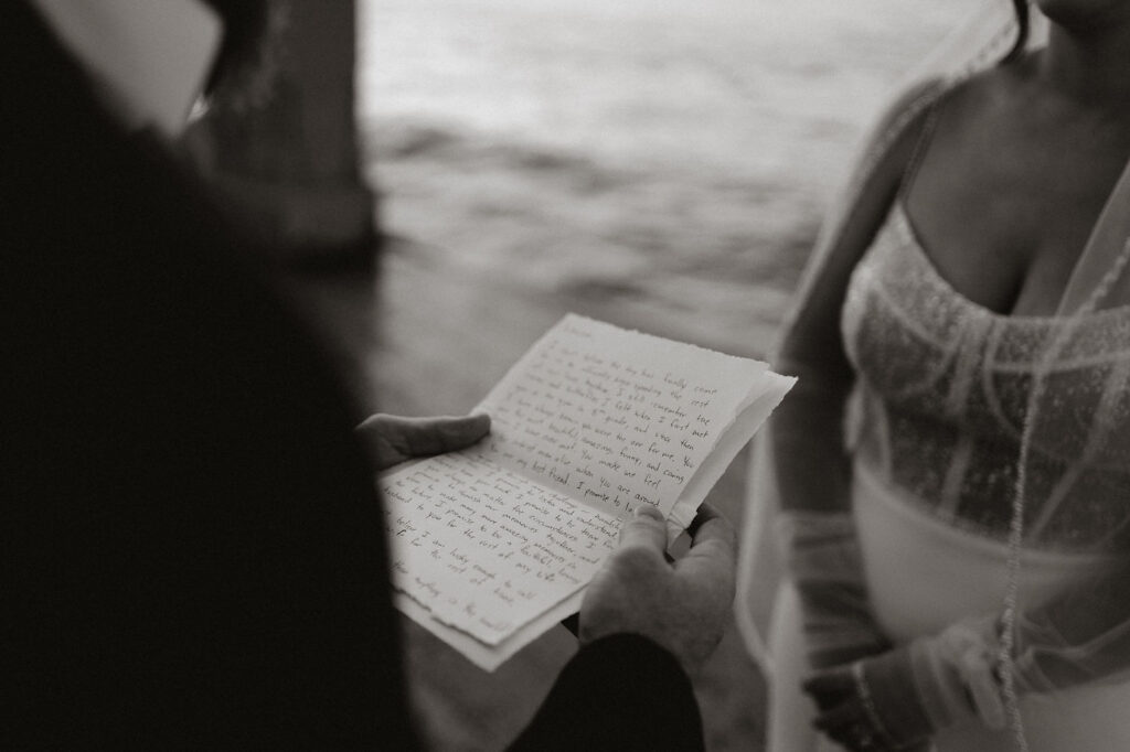 Person reading handwritten letter to woman in sparkling dress. They're standing by a waterfront. Emotional, intimate setting. Black and white.