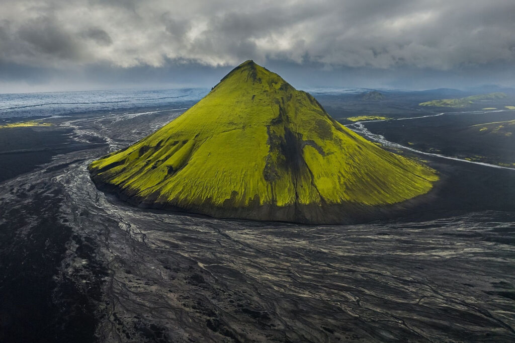 best place to elope in iceland: Fjallabak Nature Reserve. Green, moss-covered volcano in a dark, volcanic landscape under cloudy skies, with winding rivers. Moody and dramatic setting.