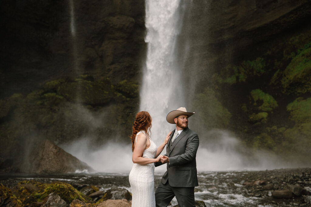 bride and groom stands near a waterfall; the man wears a cowboy hat. Lush greenery surrounds them, creating a romantic scene at the best place to elope in iceland