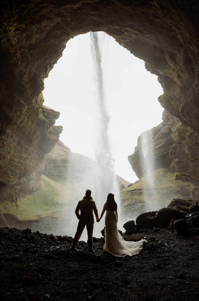 bride and groom holding hands under a cave opening with a waterfall in the background. The setting is dramatic and serene, with earthy tones at the best place to elope in iceland