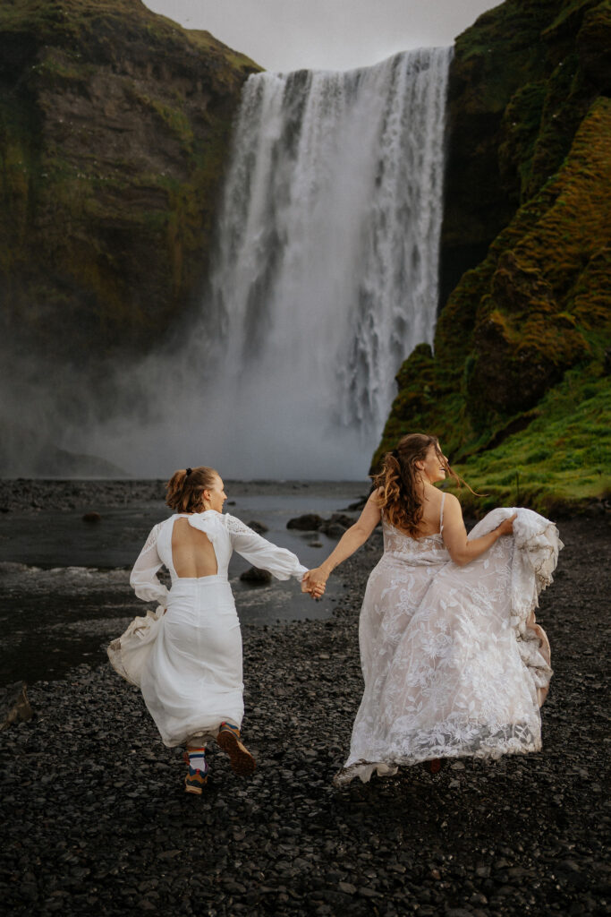 Two brides run hand in hand toward a large waterfall, set against a lush green and rocky landscape. Joyful and adventurous for an iceland elopement