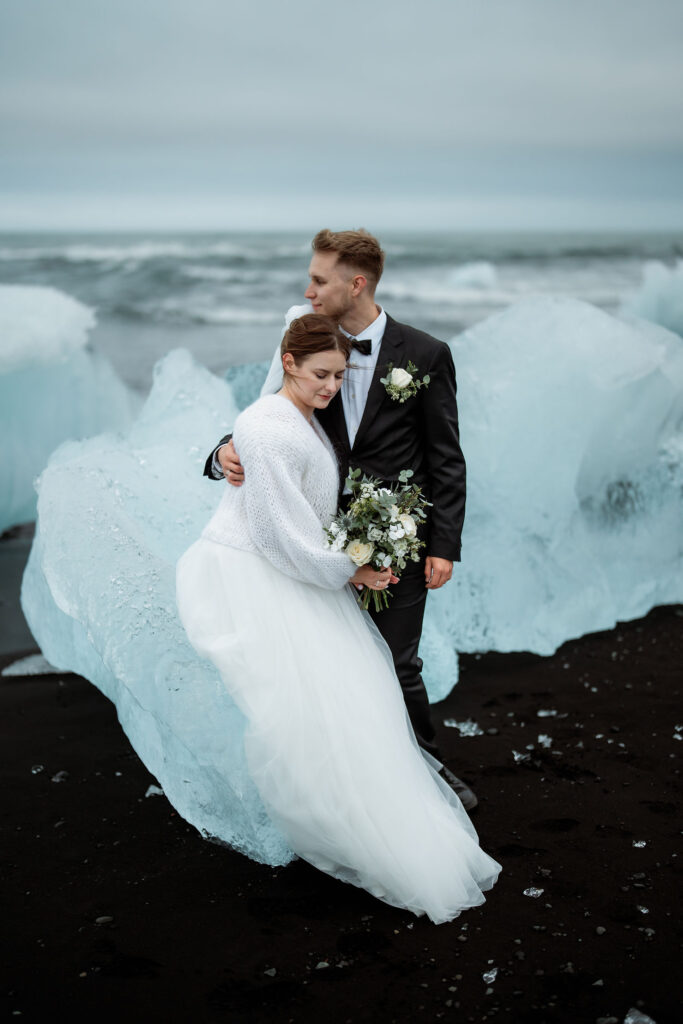 Bride and groom embrace at diamond beach in iceland with icy blue glaciers. The bride holds a bouquet, and the mood is serene and romantic.
