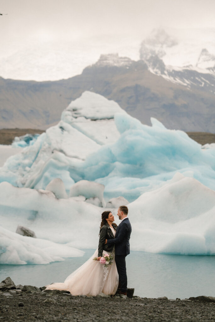 Couple embraces on rocky shore with blue icebergs in background. Woman holds bouquet, wearing a flowing elopement dress; serene, romantic atmosphere in iceland