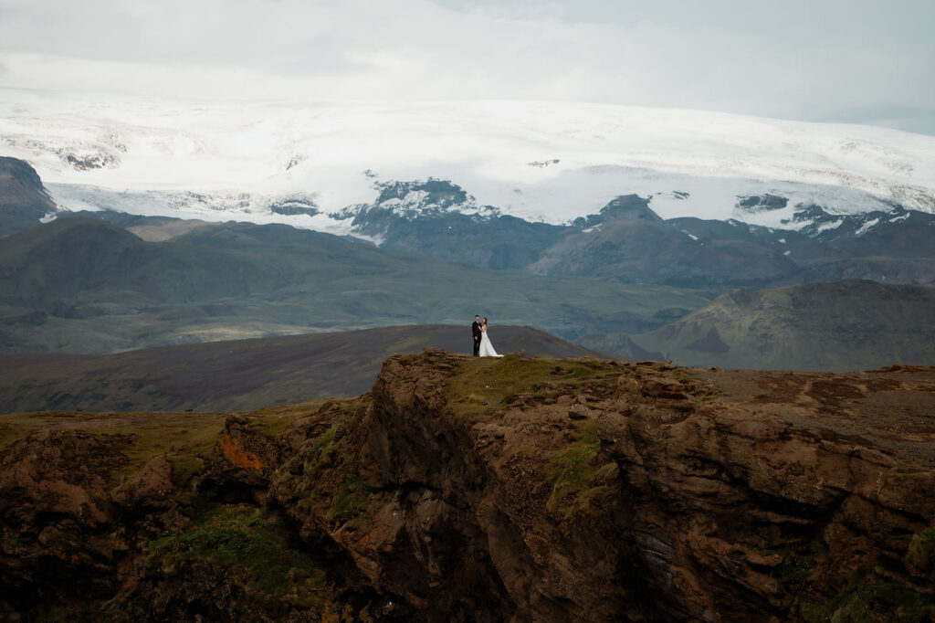 Bride and groom stand on a rocky cliff, with a vast landscape and snow-capped mountains in the background, conveying a serene mood at the best place to elope in iceland.