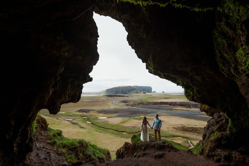 bride and groom holding hands in a cave entrance overlooking a vast, rocky landscape. Overcast sky, lush greens, and distant hills visible during iceland elopement