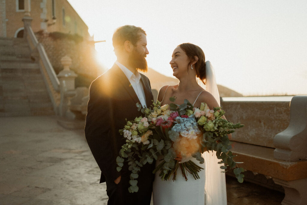 Bride and groom stand on stone steps, exchanging vows at sunset in Barcelona. Background features a historic tower, creating a romantic ambiance.
