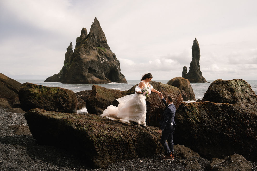 Bride and groom walk across rocky edge of balck sand beach during iceland elopement