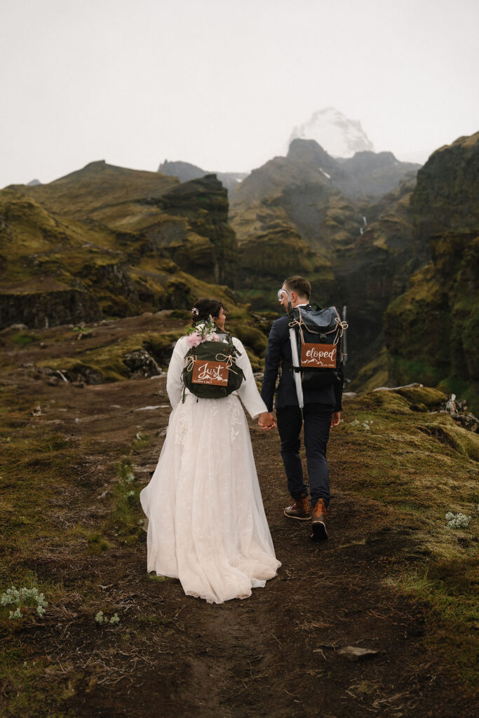 bride and groom with backpacks reading "Just Eloped" walks hand-in-hand on a mossy mountain path under a cloudy sky during iceland elopement
